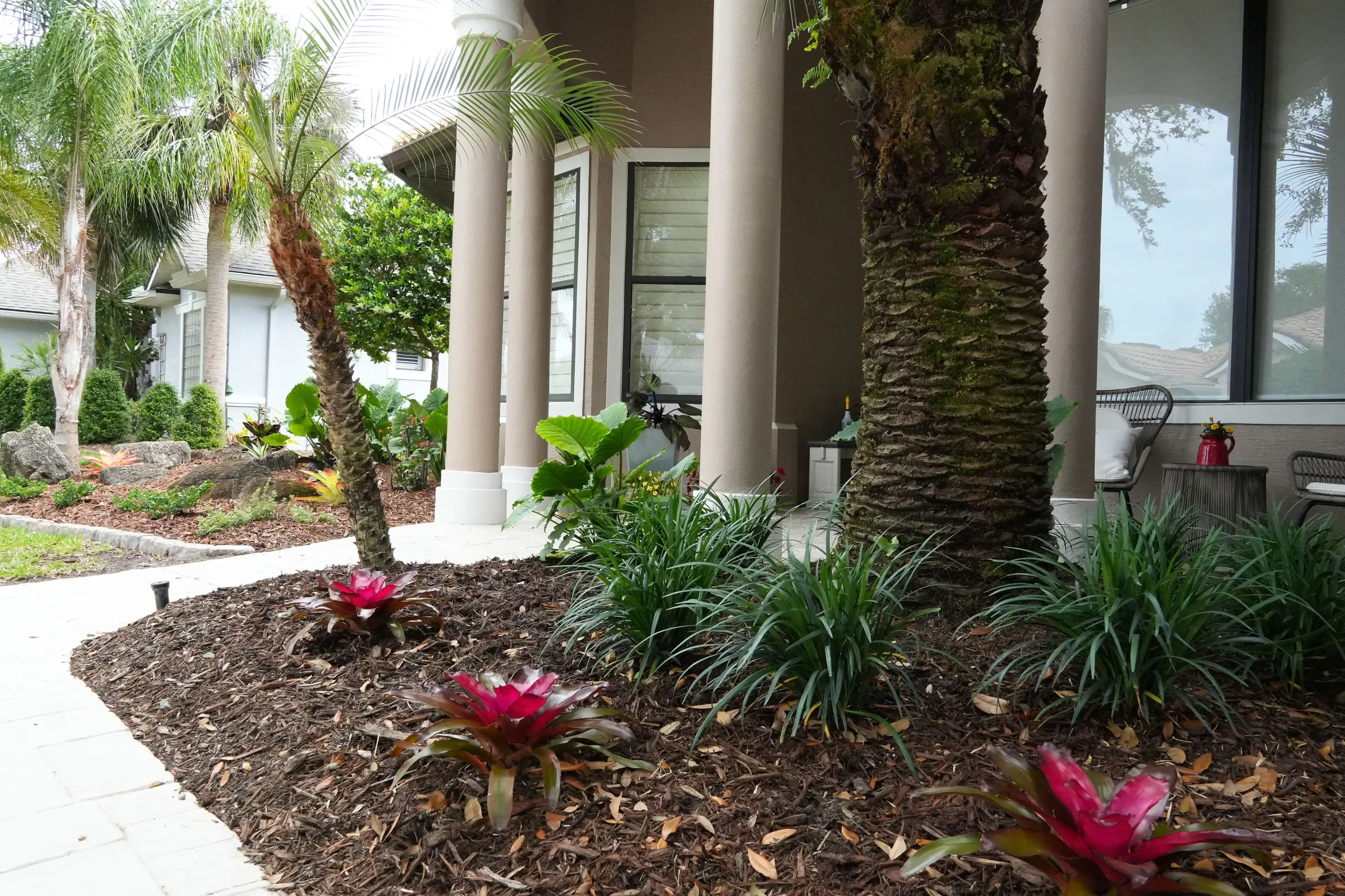 Curved mulch bed with bromeliads, liriope, and a young palm along a paver walkway to a residential entryway